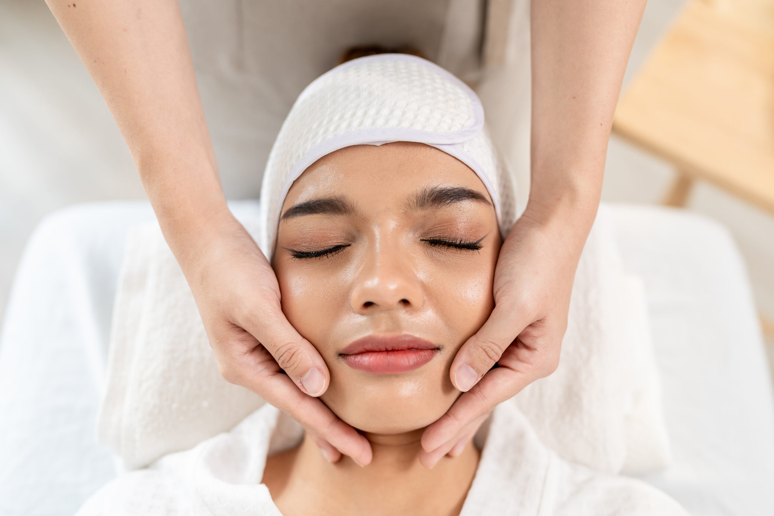 Beautiful woman getting head and facial massage treatment at beauty spa. Attractive girl lying down on massage bed, getting relaxing physiotherapy service from masseuse for body care in spa clinic.